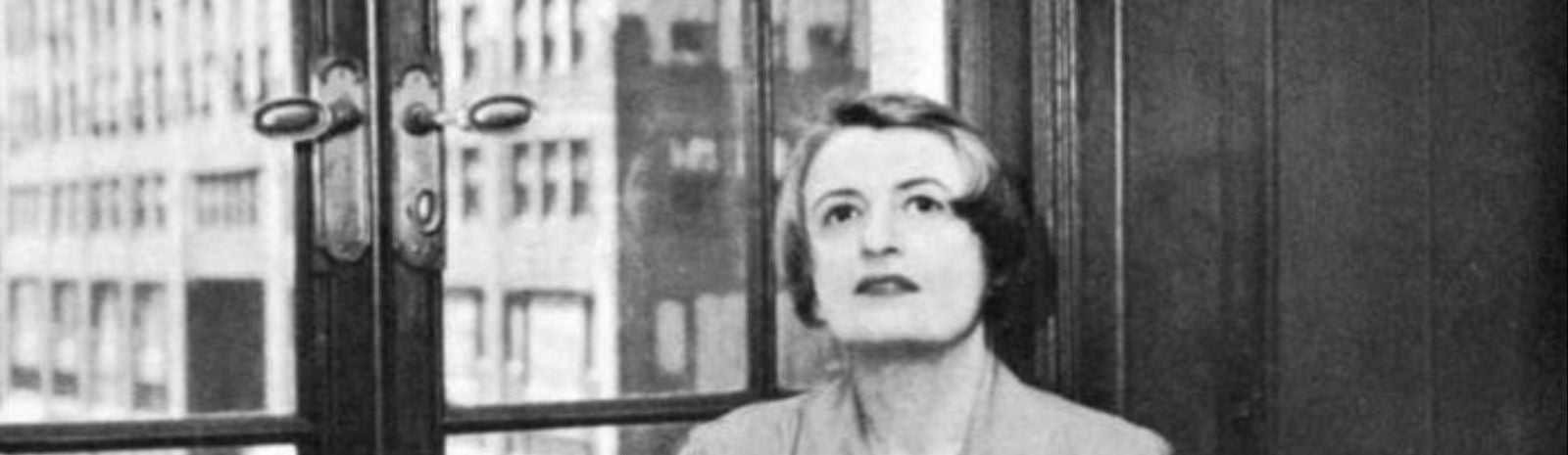 Black and white photo of Ayn Rand looking off into the distance, her hair cropped into a bob, wearing a 1950s style shirt dress by a window in a high rise apartment.