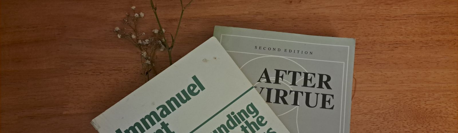 A closeup of two books, one overlapping each other. The one on the bottom of the pile is After Virtue, the one overlapping, a beaten up copy of The Grounding for the Metaphysics of morals, on a wooden table.
