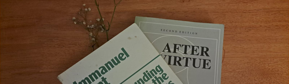 A closeup of two books, one overlapping each other. The one on the bottom of the pile is After Virtue, the one overlapping, a beaten up copy of The Grounding for the Metaphysics of morals, on a wooden table.