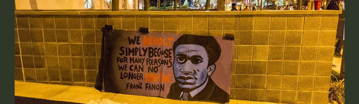 A poster with Frantz Fanon's portrait leaned against a small tiled wall. Reads: We revolt simply because, for many reasons we can no longer breathe. -Franz [sic] Fanon