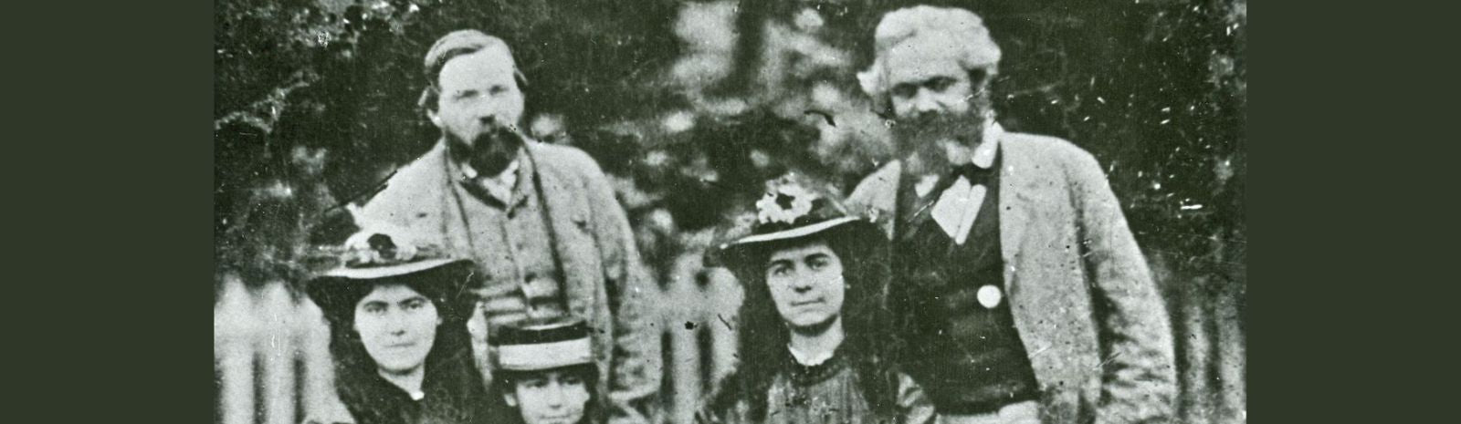 A photo of Friedrich Engels on the left, Karl Marx on the right, and three of Marx's daughters in the middle, in black and white.
